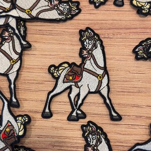 May include: Embroidered patch featuring a cartoon horse with a saddle. The horse is light gray with a brown saddle and bridle. The patch has a black border and gold accents. The horse is smiling. Patches are scattered on a wooden surface.