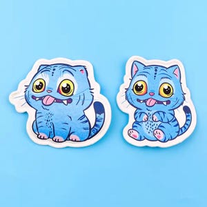 May include: Two blue cartoon cat stickers with large yellow eyes and pink tongues sticking out. The cats have blue stripes and are on a white background. The background is a solid light blue.