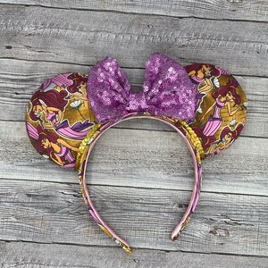 K&ouml;nnte beinhalten: Ein Stirnband mit zwei runden Ohren mit goldenem Hintergrund und einem Cartoon-Charakter-Design. Das Stirnband hat ein rosa Band mit passendem Design. Eine gro&szlig;e, paillettenbesetzte lila Schleife sitzt zwischen den Ohren.