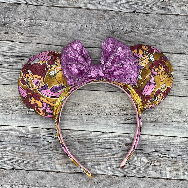 K&ouml;nnte beinhalten: Ein Stirnband mit zwei runden Ohren mit goldenem Hintergrund und einem Cartoon-Charakter-Design. Das Stirnband hat ein rosa Band mit passendem Design. Eine gro&szlig;e, paillettenbesetzte lila Schleife sitzt zwischen den Ohren.
