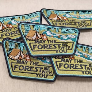 May include: Embroidered patches with a camping theme. Each patch features a tent, campfire, and the text "MAY THE FOREST BE WITH YOU" in a rectangular frame. The patches have a green, blue, and brown color scheme.