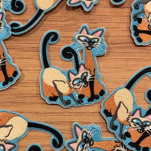 Może przedstawiać: Haftowane naszywki przedstawiające koty syjamskie w zabawnej pozie. Naszywki mają jasnoniebieską obwódkę, z kotami w kolorze beżowym, pomarańczowym i czarnym. Koty mają niebieskie oczy i różowe uszy. Naszywki są na drewnianej powierzchni.