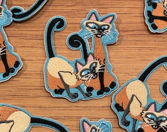 May include: Embroidered patches featuring Siamese cats in a playful pose. The patches have a light blue border, with the cats in tan, orange, and black. The cats have blue eyes and pink ears. The patches are on a wooden surface.