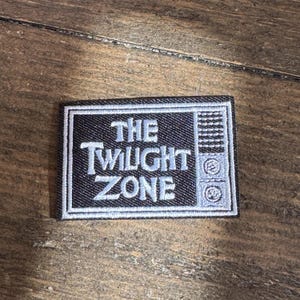Peut inclure: Un patch brodé noir et blanc avec le texte "The Twilight Zone" dans une police de caractères stylisée. Le patch est en forme d'écran de télévision avec une bordure blanche.