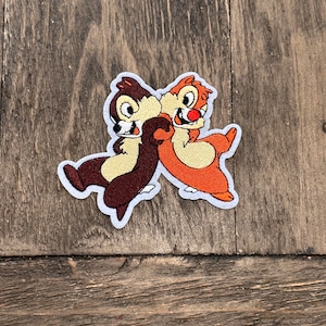 May include: Embroidered patch featuring Chip and Dale from Disney's Chip 'n Dale Rescue Rangers. The patch shows the two chipmunks in a playful pose, with Chip on the left and Dale on the right. The patch is made of felt and has a white background.