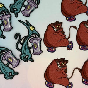 Könnte beinhalten: Gestickte Aufnäher mit Zeichentrickfiguren. Ein Aufnäher zeigt eine türkisfarbene Kreatur, die einen lila Behälter hält. Ein weiterer Aufnäher zeigt eine kastanienbraune Figur mit Schwanz und Turnschuhen. Die Aufnäher haben detaillierte Nähte und lebendige Farben.
