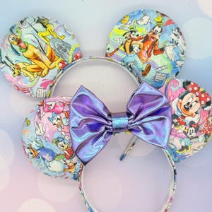 Puede incluir: Un conjunto de diademas con orejas de ratón con un gran lazo morado iridiscente. Las orejas están decoradas con personajes de dibujos animados coloridos, incluyendo a Pluto, Goofy, Donald Duck y Minnie Mouse, sobre un fondo azul claro.