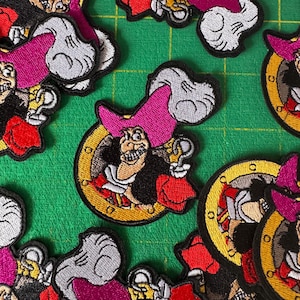 May include: Embroidered patches featuring Captain Hook from Peter Pan. The patches showcase the character's face, a pink hat, and a red coat, set against a gold-colored clock face. The patches are on a green surface.