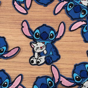 May include: Embroidered patches featuring the blue alien Stitch from Lilo & Stitch, hugging a white kitten. The patches have pink ears and accents, with black and white details. These patches are ideal for decorating clothing or accessories.