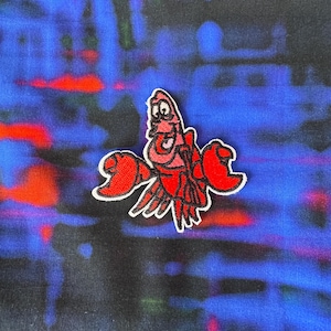 May include: Red and pink cartoon lobster with a smiling face and large claws. The lobster is embroidered on a blue and red fabric.