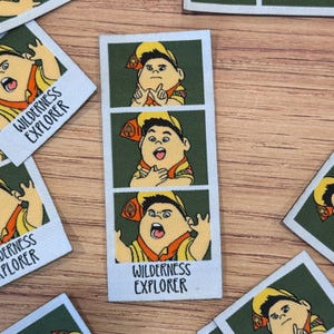 May include: Fabric patches featuring a cartoon character in a Wilderness Explorer uniform. The character is depicted in three different poses within a green frame, with the words "Wilderness Explorer" printed below. The patches are on a wooden surface.