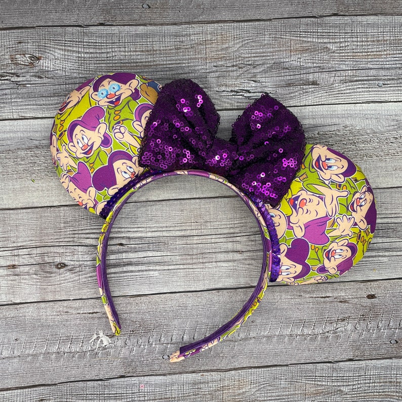 Pu&ograve; includere: Orecchie di topo viola e verdi con un fiocco di paillettes viola. Le orecchie presentano un motivo ripetitivo dei sette nani del film Disney Biancaneve e i sette nani.