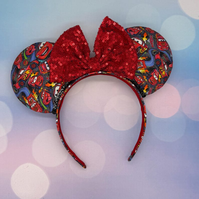 May include: Headband with large, red sequined bow and matching ears. The ears are patterned with images of the Cars movie characters. The headband is red with a Cars-themed pattern. The ears are approximately 4 inches in diameter.
