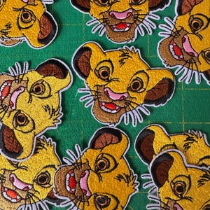 Puede incluir: Parches bordados con la cara de un cachorro de león sonriente con detalles amarillos, marrones y rosas. Los parches tienen bigotes de hilo blanco y están dispuestos sobre una superficie verde. El cachorro de león tiene una expresión feliz.