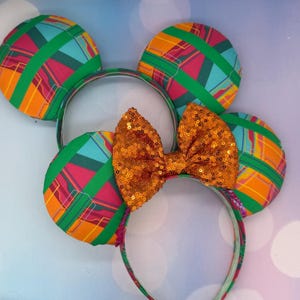 May include: A pair of headband ears featuring a vibrant geometric pattern in shades of orange, teal, magenta, and green. The ears are accented with a central, shimmering orange sequined bow. The headband frame is covered in a matching fabric.