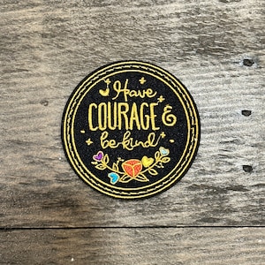 Op de afbeelding: Een ronde, zwarte patch met gouden rand en de gouden tekst "I Have COURAGE & be kind". De patch heeft kleurrijke bloemen- en vlindermotieven, tegen een houten achtergrond.