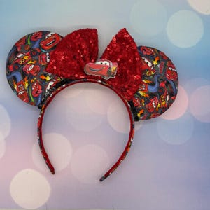 May include: A Cars-themed headband featuring two round ears patterned with race cars and roads. A large, red sequined bow sits between the ears, with a small Lightning McQueen emblem. The headband itself is red.