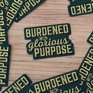 May include: Dark green embroidered patches with gold lettering. The patches read "BURDENED with glorious PURPOSE". The patches are rectangular with a unique shape and are scattered on a wooden surface.