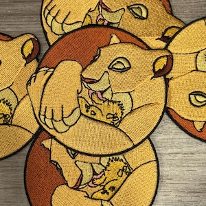 May include: Embroidered patches featuring a lioness and cub in shades of tan, brown, and pink. The patches are circular and show the lioness embracing the cub. The patches are likely for clothing or accessories.