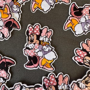 Pode incluir: Patches bordados com Minnie Mouse e Margarida em várias poses. Os patches têm uma borda branca e mostram os personagens em rosa, amarelo e preto. Os patches estão espalhados em uma superfície cinza escuro.