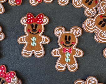 May include: Brown gingerbread man-shaped felt ornaments with white icing details. One has a red bow with white polka dots, the other a red bow tie. Both have green and white accents. Festive holiday decor.