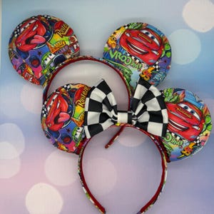 May include: A set of themed mouse ears featuring Lightning McQueen from the movie Cars. The ears have a colorful design with the character's image, the text "VROOOM!" and a checkered bow. The headband is red.