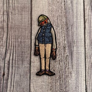 May include: A brown and beige embroidered patch of a person wearing a blue vest, khaki pants, a green hat, and orange sunglasses.