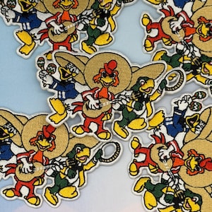 May include: Embroidered patches featuring cartoon chickens wearing sombreros and playing musical instruments. The chickens are in various colors, including red, green, and blue, with yellow accents. The patches are on a light blue background.