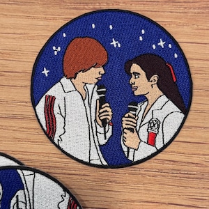 May include: Embroidered patch featuring two people singing into microphones, set against a dark blue background with white stars. The figures are wearing white jackets with red accents. The patch is circular and has a black border.