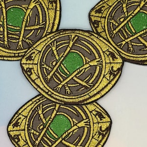 May include: Embroidered patches with a gold-coloured, eye-shaped design, brown background, and green centre. The patches have a detailed, stitched appearance, with multiple patches visible.