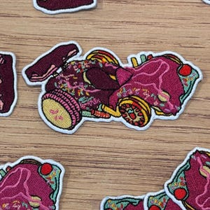 Pode incluir: Patch bordado com um design estilizado de carro de corrida. O patch é principalmente bordô com detalhes amarelos e verdes e uma borda branca. O carro tem um spoiler e rodas detalhadas. O patch é provavelmente para roupas ou acessórios.