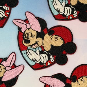 May include: Embroidered patches featuring Minnie and Mickey Mouse in a heart-shaped design. Minnie wears a pink bow and Mickey has a red hat. The patches are primarily red, black, and pink, with light blue accents. These patches are suitable for clothing or accessories.