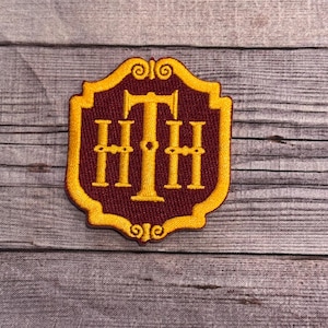 May include: A gold embroidered patch featuring the letters "HHH" on a burgundy background. The patch is shaped like a shield with a decorative top.
