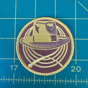 May include: A round, embroidered patch with a brown background and a gold outline. The patch features a brown hat with a gold brim and a gold rope design.
