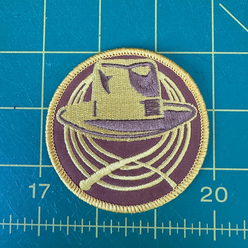 May include: A round, embroidered patch with a brown background and a gold outline. The patch features a brown hat with a gold brim and a gold rope design.