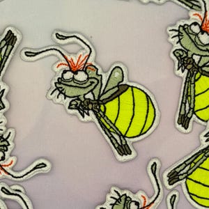 May include: Embroidered patches featuring a cartoon firefly design. The fireflies have a green head, yellow abdomen, and brown wings. The patches are outlined in white and have orange accents on the head.
