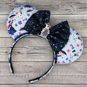 Puede incluir: Un par de orejas de Minnie Mouse de tela azul y blanca con un lazo de lentejuelas negras. Las orejas presentan un patrón de pingüinos, flores y un personaje de la película 'Cruella'.