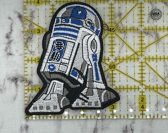 May include: A white and blue embroidered patch of R2-D2, a robot character from the Star Wars franchise. The patch is made in the USA.