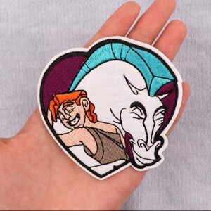 Könnte beinhalten: Gebügelter Aufnäher mit gesticktem Cartoon-Bild von Herkules und Pegasus aus dem Disney-Film Herkules. Der Aufnäher hat die Form eines Herzens und ist in den Farben Lila, Weiß und Blau gehalten.