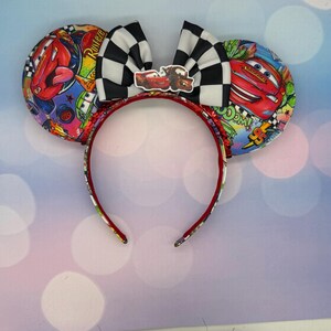 May include: A Cars-themed headband featuring large, round ears with colorful graphics of Lightning McQueen and other characters. A black and white checkered bow sits atop the headband, with a small Cars character emblem. The headband is red with a patterned design.