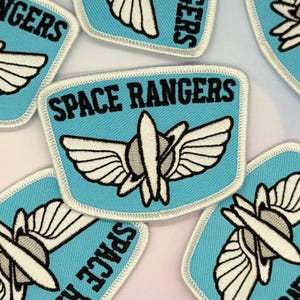 Può includere: Un gruppo di toppe ricamate azzurre con la scritta "SPACE RANGERS" in nero. Ogni toppa presenta un disegno stilizzato bianco e grigio di un'astronave alata. Le toppe hanno un bordo bianco.