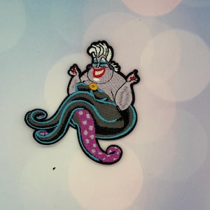 Op de afbeelding: Een geborduurde strijkpatch met een cartoon-zeeheks met wit haar, een bleke huid en een brede glimlach met rode lippen. Het figuur heeft zwarte inktvisarmen met paarse, gespikkelde onderkanten en blauwe omlijningen, en draagt een zwarte jurk.