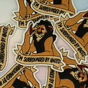 Op de afbeelding: Geborduurde patch met een cartoonleeuw met een zwarte manen en een banner met de tekst "I'M SURROUNDED BY IDIOTS". De leeuw bevindt zich in een benarde pose, met zijn mond open. De patch heeft een bruine achtergrond en een witte rand.