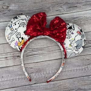 Può includere: Fascia per capelli con fiocco di paillettes rosse e orecchie bianche con stampa di cani dei cartoni animati. Le orecchie hanno uno sfondo bianco con cani dei cartoni animati neri e rossi, ossa e impronte di zampe.