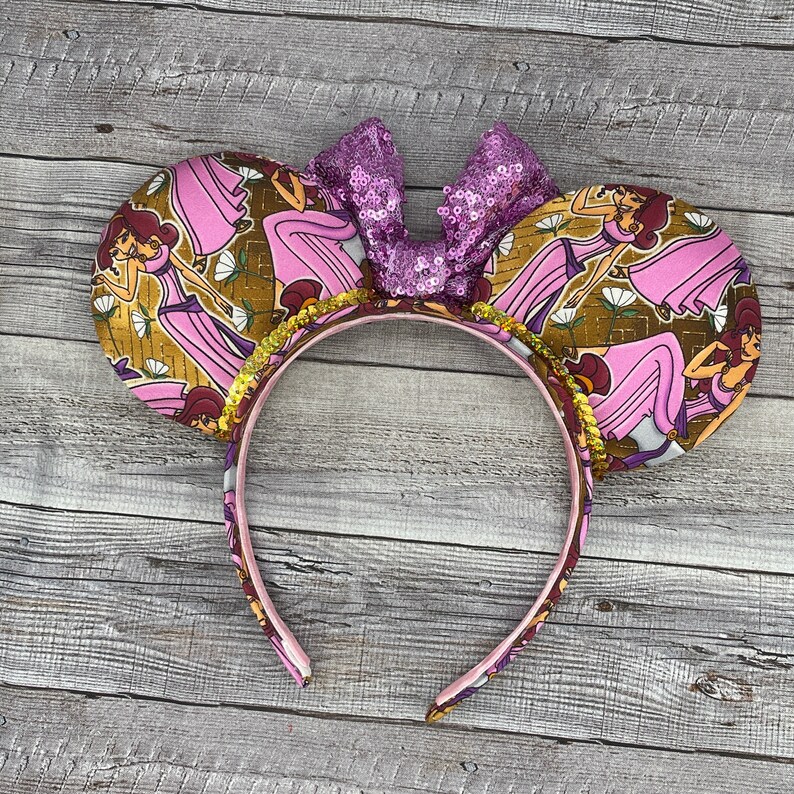 K&ouml;nnte beinhalten: Ein Stirnband mit Mauseohren mit einem rosa-goldenen Muster mit einer Cartoonfigur. Die Ohren sind mit einer lila Pailletten-Schleife und goldenen Verzierungen versehen. Das Stirnband ist rosa und gemustert.