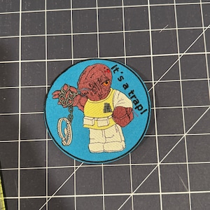 May include: A blue and white embroidered patch featuring a cartoon character with a chain and the text "It's a trap!"