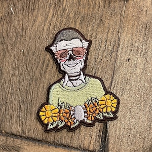 May include: Embroidered patch featuring a cartoon character wearing sunglasses and a green sweater, surrounded by yellow flowers.
