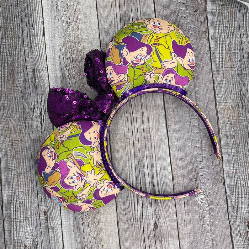 Pu&ograve; includere: Orecchie di Minnie Mouse viola e verdi con un fiocco di paillettes viola e una stampa dei sette nani di Biancaneve.