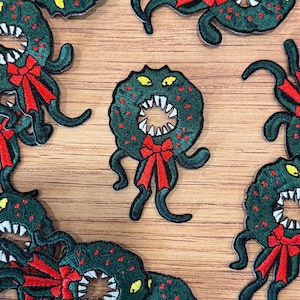Könnte beinhalten: Gestickte grüne Kranzaufnäher mit roten Schleifenakzenten und tentakelartigen Anhängseln. Die Kränze haben weiße Zähne und gelbe Augen. Die Aufnäher sind auf einer hellbraunen Holzoberfläche verstreut.