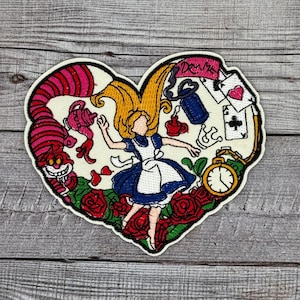 Könnte beinhalten: Gestickter herzförmiger Patch mit Alice aus Alice im Wunderland, die in ein Kaninchenloch fällt. Der Patch enthält eine Grinsekatze, Spielkarten, eine Taschenuhr und Rosen.